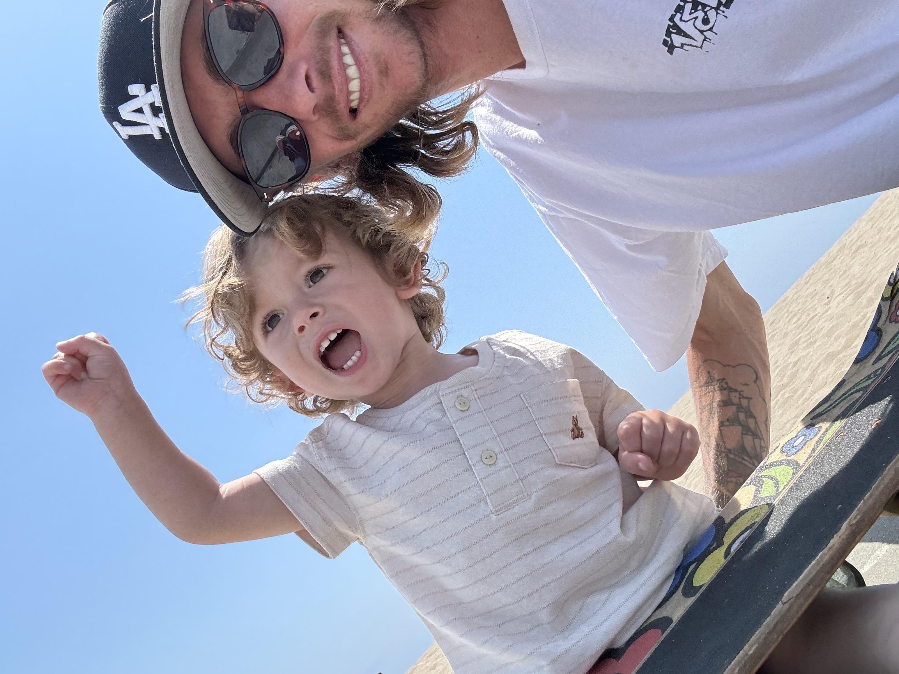 Haden smiling with a young child on a skateboard under a bright blue sky.